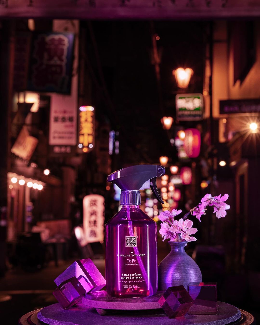Home perfume spray by Rituals stands on a round pedestal with decorative purple crystal shapes in a Japanese-style, dark, urban setting.