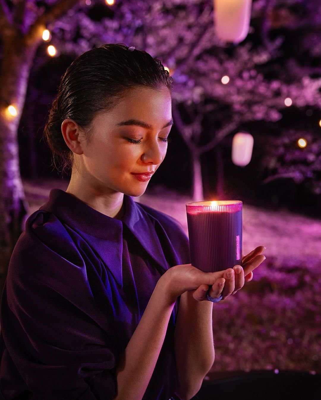 Woman holding an illuminated purple candle outdoors at night. The scene is set with trees and string lights for a warm, ambient atmosphere.