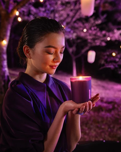 Woman holding an illuminated purple candle outdoors at night. The scene is set with trees and string lights for a warm, ambient atmosphere.