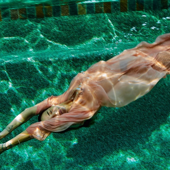Underwater view of a woman in a peach dress diving into a swimming pool with turquoise water, creating a serene and ethereal scene.