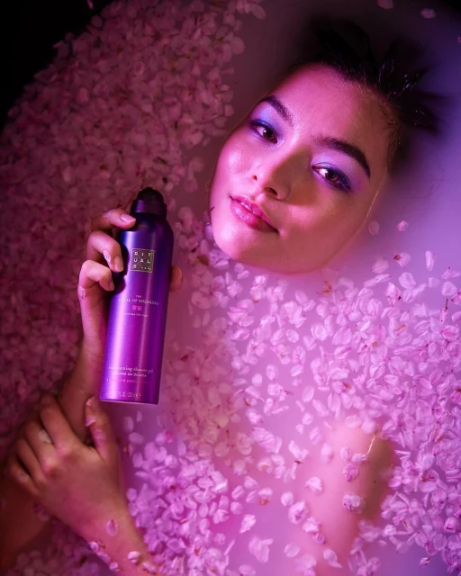 Beautiful young woman relaxing in a milky bath with cherry blossom petals, holding a purple bottle of Rituals shower oil, beauty concept.