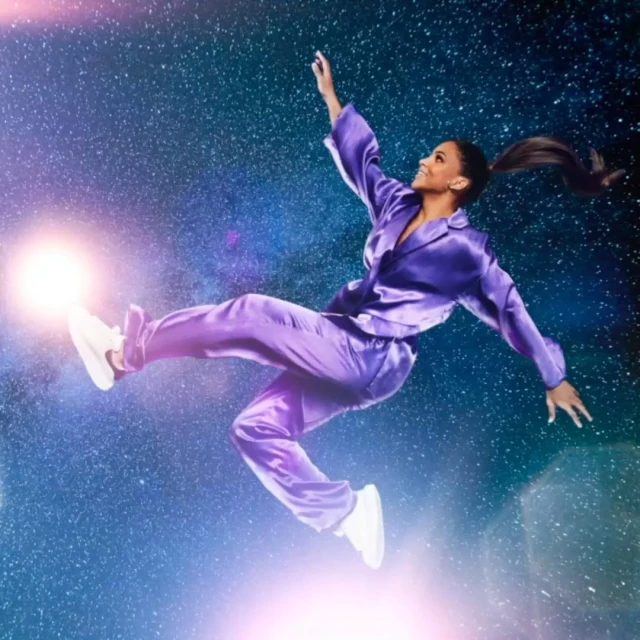 Full shot of a woman in a purple pajama set floating in space with legs extended and one arm raised, smiling in a lighthearted image.