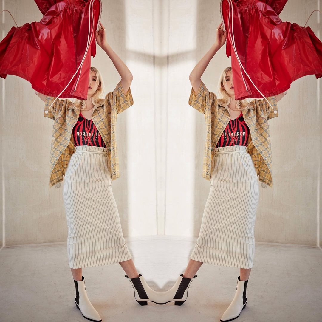 Young woman in a patterned blouse and white skirt raises a red garment overhead indoors with a mirrored background, full body shot.
