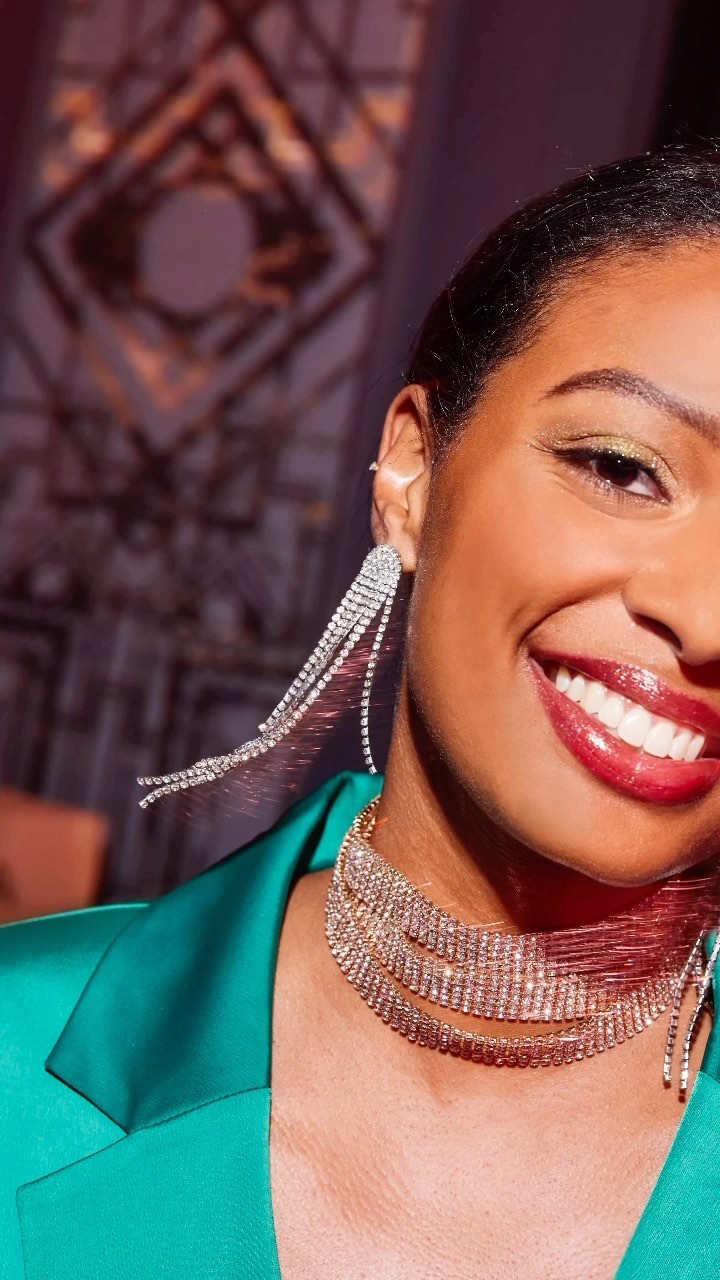 Close-up of a smiling adult female with diamond jewelry including earrings and necklace, wearing green jacket, against dark background.