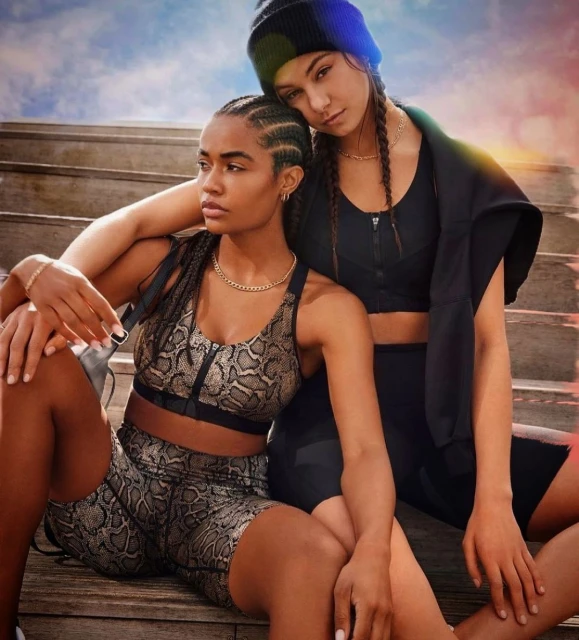 Two women in activewear sit on steps. One wears a snake print bra and shorts. The other wears black activewear with a beanie.