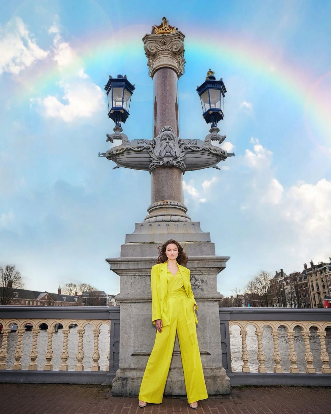 Woman in vibrant yellow suit poses on a bridge with ornate lamp post against a rainbow sky in an urban setting with waterscape.