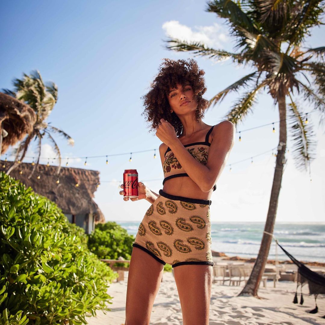 Woman in patterned two-piece holds a Coca-Cola can on a sunny beach with palm trees and a thatched-roof structure.