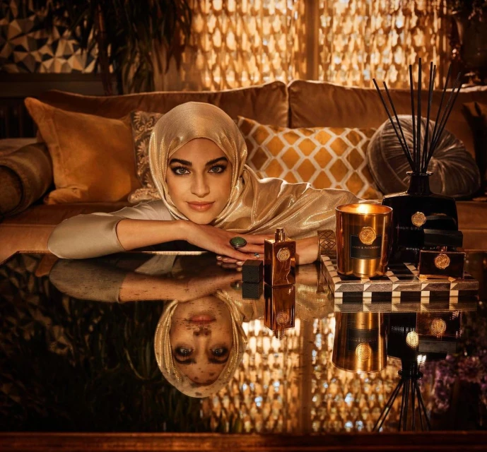 Woman in gold headscarf poses with perfume and candle collection on reflective table indoors, surrounded by pillows and textured wall.