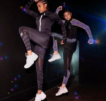 Two women in black athletic wear pose dynamically against a dark backdrop, showcasing their fitness apparel in a studio environment.