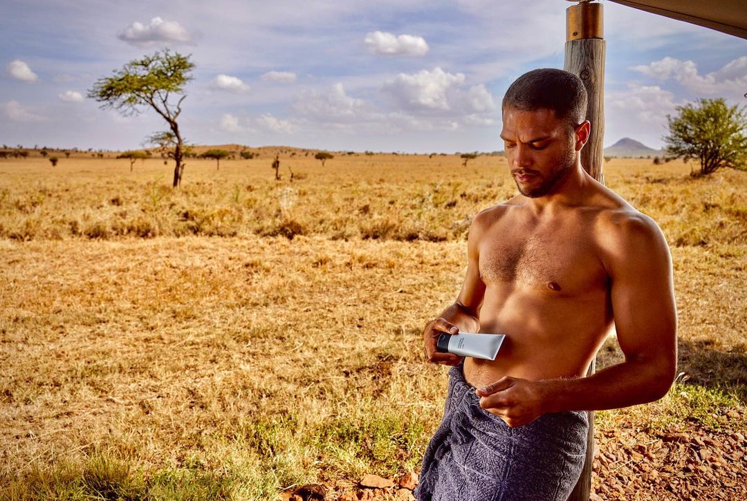 Adult male with a towel around his waist holding a skincare product in an outdoor savanna setting with trees and cloudy skies.