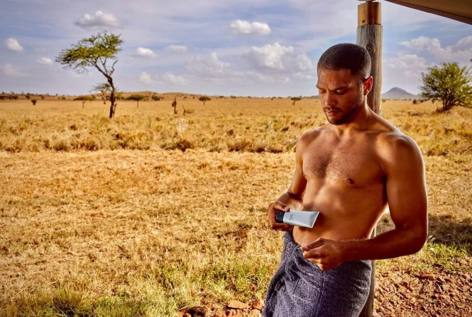 Adult male with a towel around his waist holding a skincare product in an outdoor savanna setting with trees and cloudy skies.