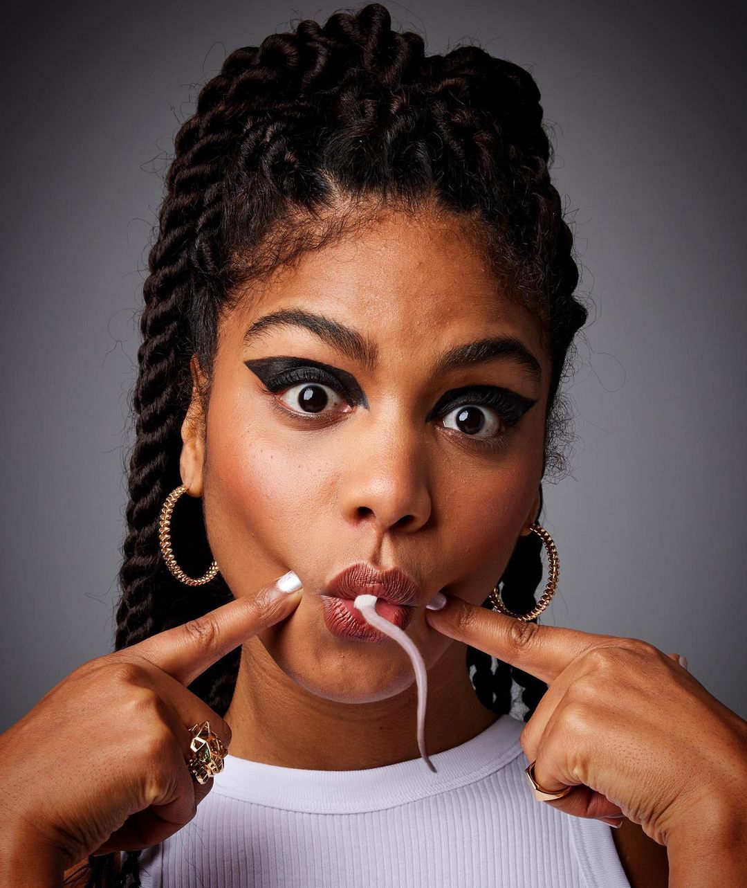 Portrait of a young woman with braided hair and heavy eyeliner, making a funny face with her fingers and a strand of saliva visible.