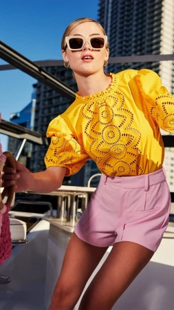 Woman on yacht in yellow eyelet top and pink shorts, sunglasses, city background, nautical setting, fashion, summer day.