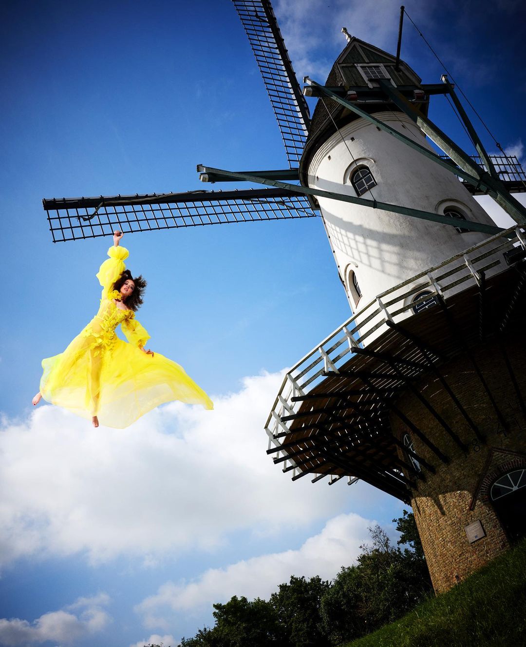 Adult woman in yellow dress jumps in front of white windmill against blue sky with few clouds on sunny day in an outdoor setting.