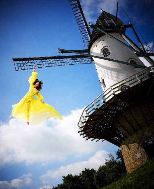 Adult woman in yellow dress jumps in front of white windmill against blue sky with few clouds on sunny day in an outdoor setting.
