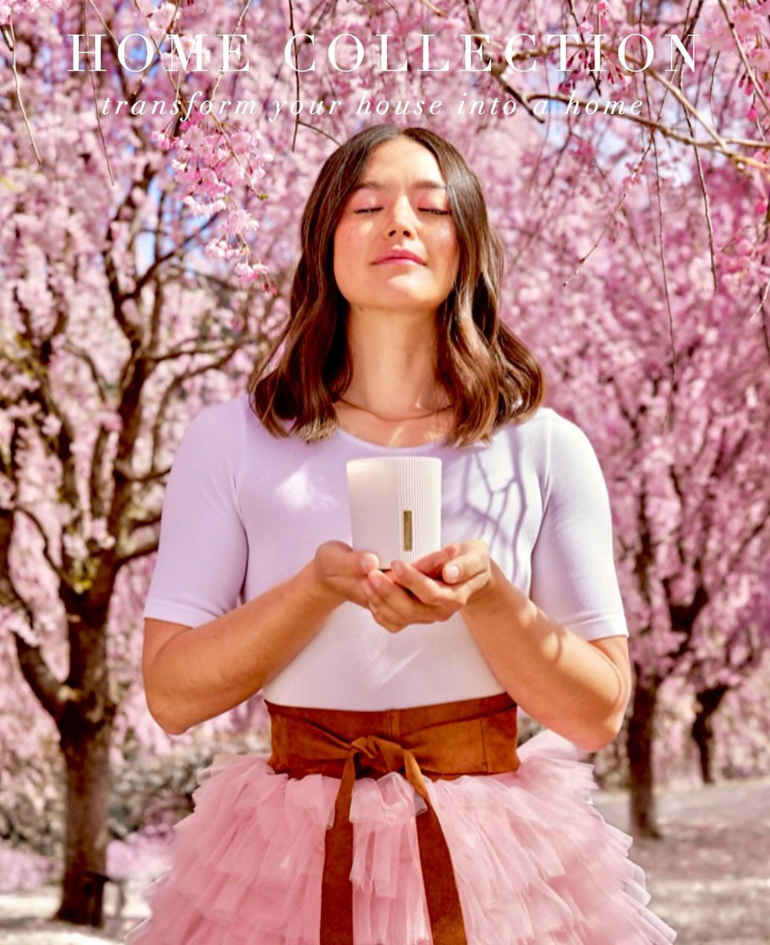 Woman with eyes closed holds a candle against a backdrop of pink cherry blossoms, wearing a white top and pink tutu skirt.