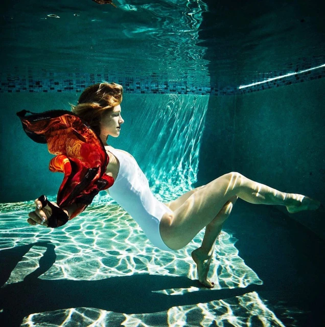 Underwater shot of a woman in a white swimsuit with an orange scarf in a swimming pool, with light patterns on the pool floor.