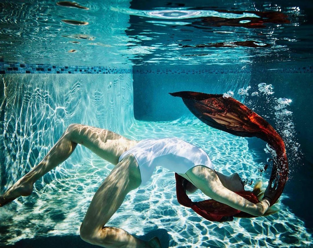 Underwater view of a woman in a white swimsuit and red fabric tail, swimming in a pool with bubbles and sunlight creating patterns.