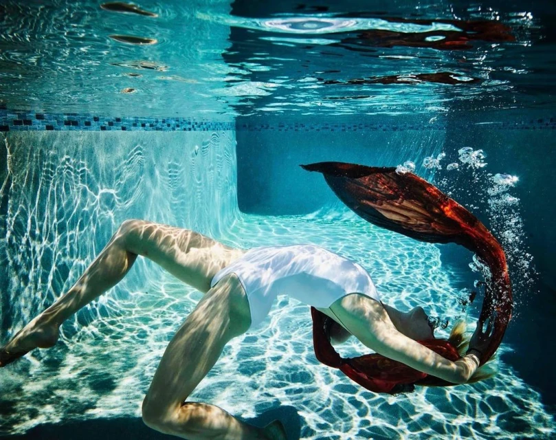Underwater view of a woman in a white swimsuit and red fabric tail, swimming in a pool with bubbles and sunlight creating patterns.