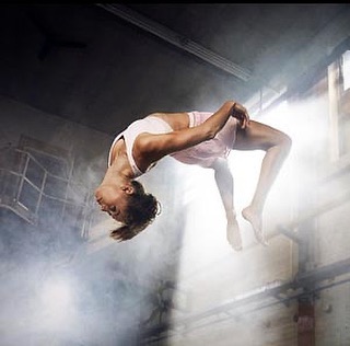 A woman in mid-air performing a backflip in a smoky, dimly lit space, wearing athletic attire, showcasing fitness and gymnastics skills.