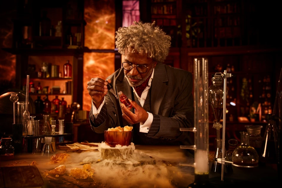 A man in a suit and glasses holds a dropper with a bottle, standing at a table filled with laboratory glassware in a dimly lit room.