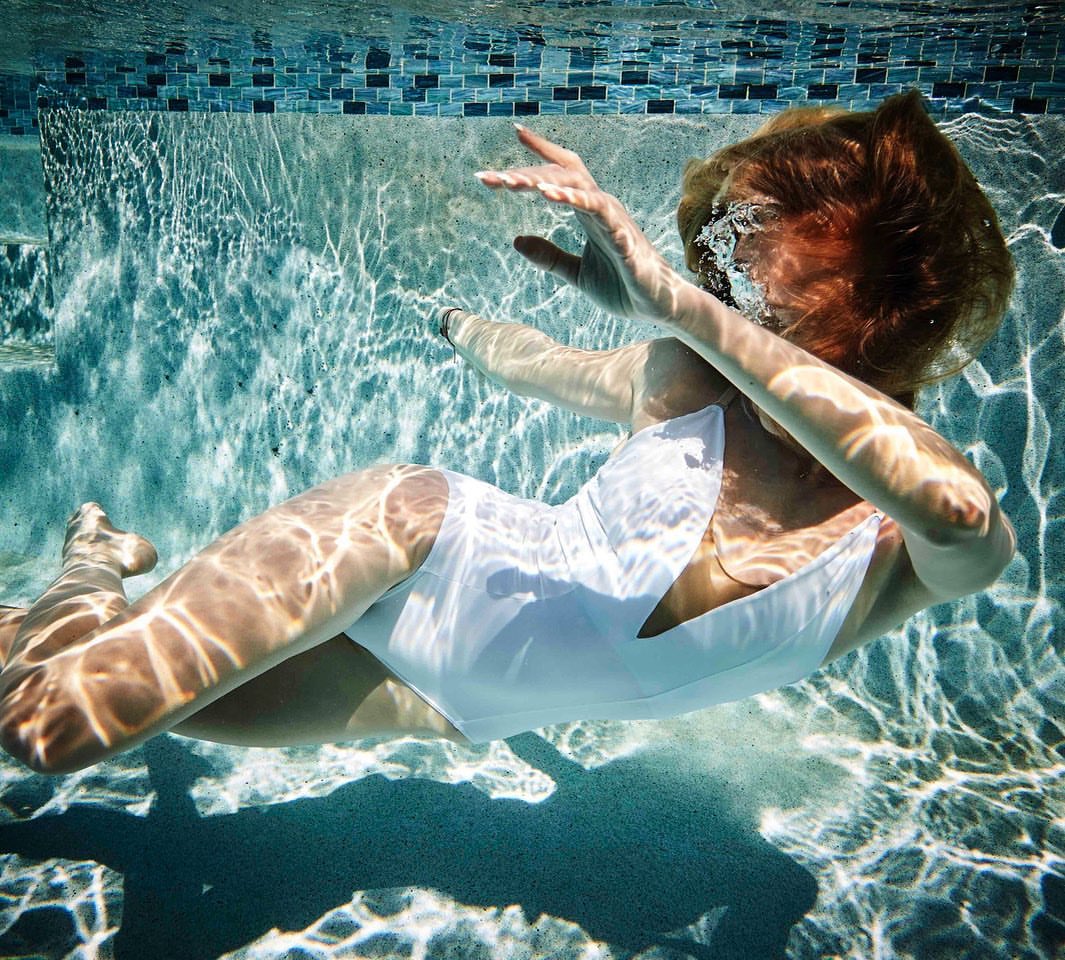 Underwater shot of a woman in a white bathing suit with diffused sunlight patterns visible on the pool's surface and blue tile border.