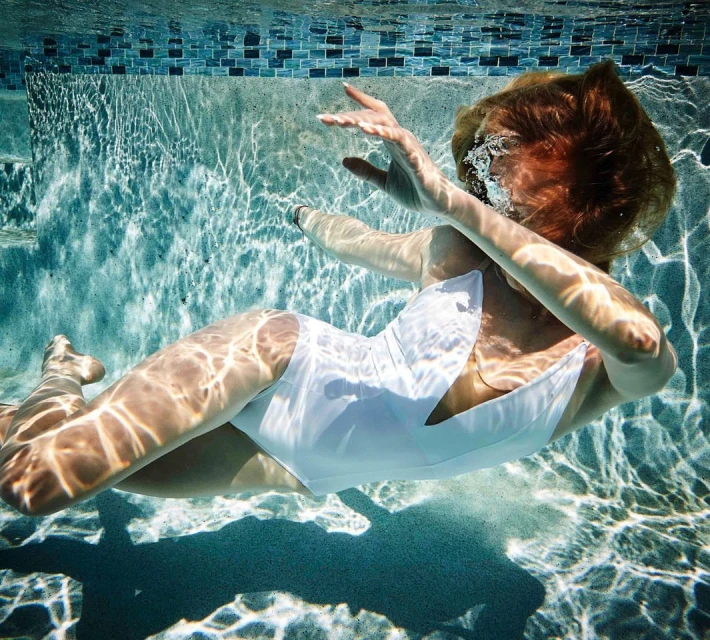 Underwater shot of a woman in a white bathing suit with diffused sunlight patterns visible on the pool's surface and blue tile border.