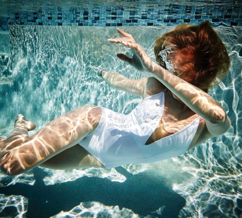 Underwater shot of a woman in a white bathing suit with diffused sunlight patterns visible on the pool's surface and blue tile border.