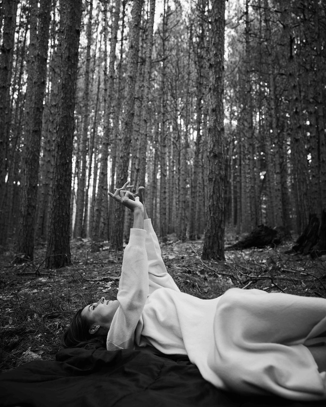Monochrome shot of a woman lying on a blanket in a forest, reaching her hands up toward the tall trees in an artistic pose.