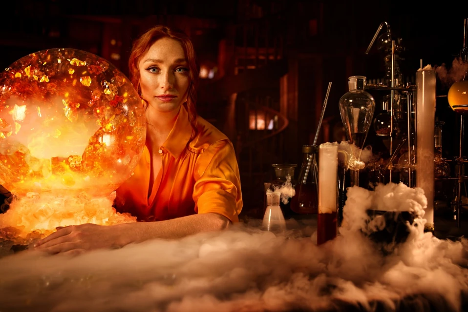 Woman with red hair poses in a vintage lab, an illuminated crystal ball in front of her amid beakers and laboratory glassware.