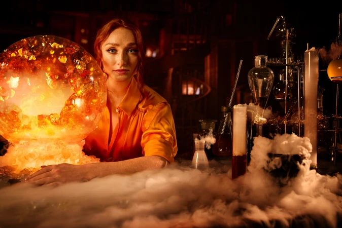 Woman with red hair poses in a vintage lab, an illuminated crystal ball in front of her amid beakers and laboratory glassware.