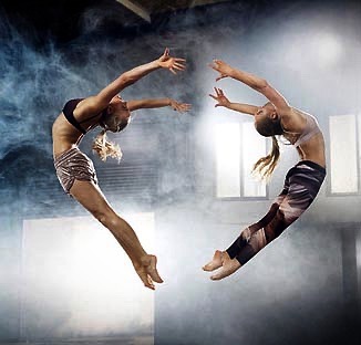 Two women are captured mid-air in a dynamic pose, wearing athletic wear, set against a backdrop of smoke or haze, creating a sense of motion.