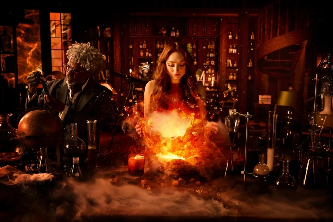 Two people, one male and one female, in a dimly lit room with shelves of bottles and a table with beakers and flasks. The woman is surrounded by an orange glow.