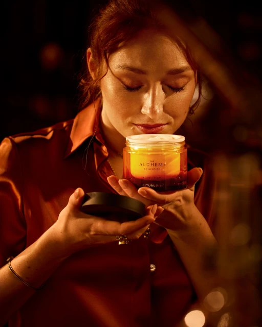 Woman with auburn hair smelling an amber-colored jar of beauty cream in dim light, wearing an orange satin shirt and silver bracelet.