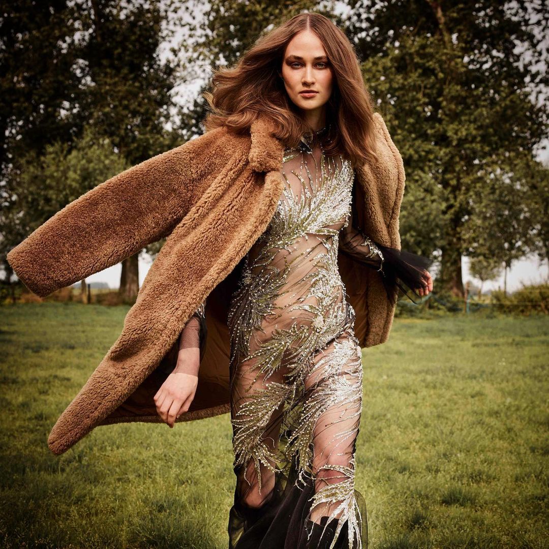 A woman with long brown hair wears a sheer dress with foliage beading, paired with a large tan fur coat, standing outdoors in a field.