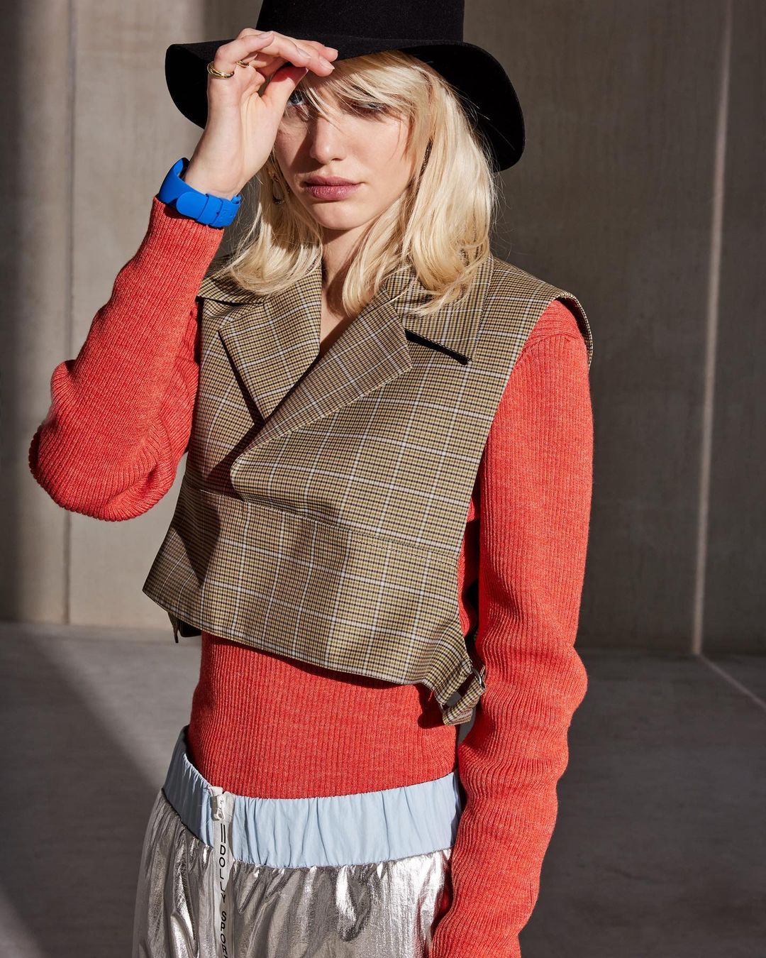 Young woman in hat and vest poses indoors against grey background, wearing orange top and metallic pants in a fashion shot.