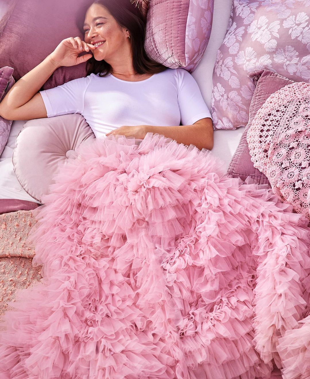 Smiling woman lies on bed surrounded by mauve pillows, under a massive pink ruffled tulle blanket, looking left with hand near mouth.