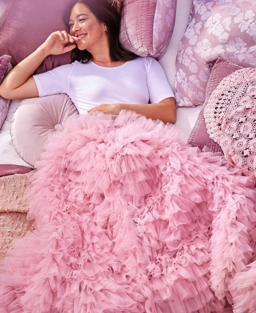 Smiling woman lies on bed surrounded by mauve pillows, under a massive pink ruffled tulle blanket, looking left with hand near mouth.