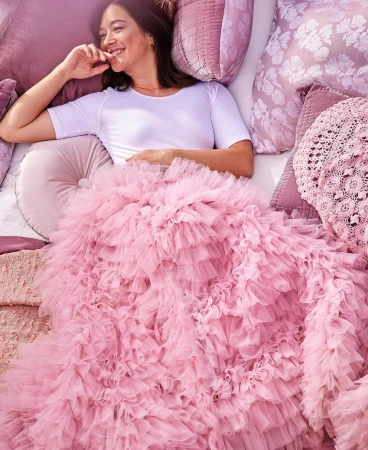 Smiling woman lies on bed surrounded by mauve pillows, under a massive pink ruffled tulle blanket, looking left with hand near mouth.