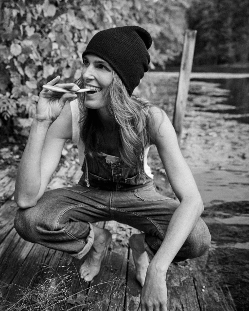 Black and white portrait of adult woman in beanie squatting on wood planks, looking up, and holding a cigarette in her mouth.