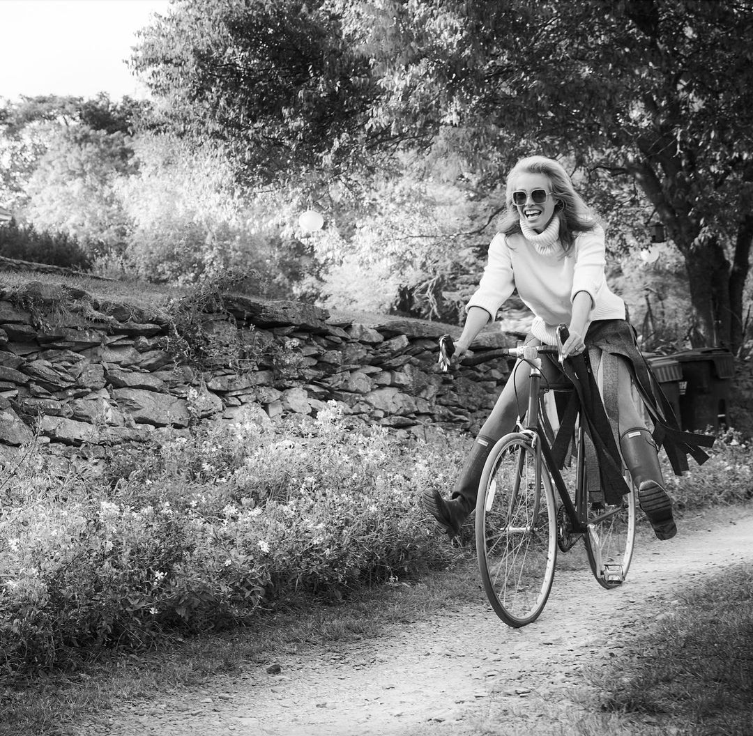 Black and white photo of a smiling woman riding a bicycle on a dirt path in a rural setting with trees and a stone wall in the background.