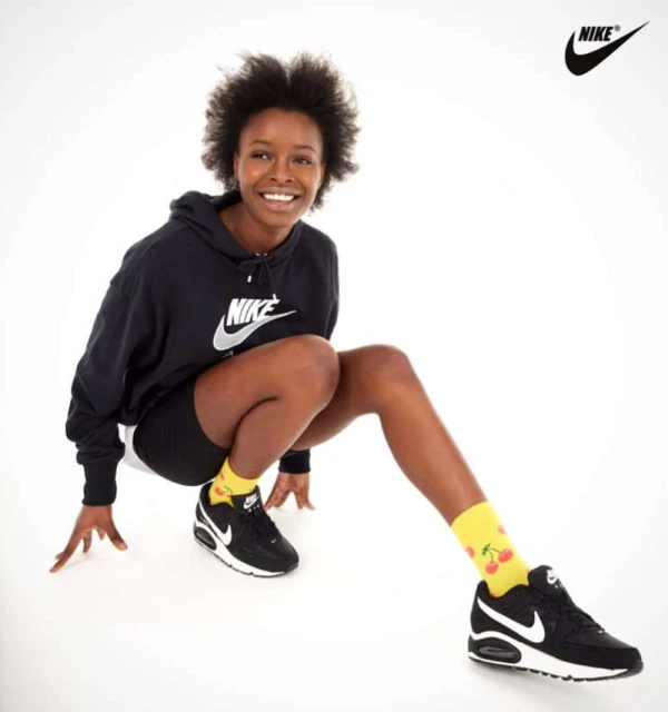 Smiling woman poses in black Nike hoodie and bike shorts with black Nike sneakers and cherry-print socks against a white backdrop.