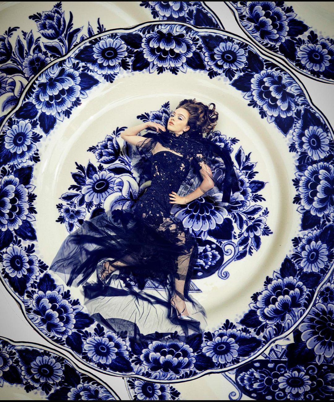 Woman in an elaborate dark couture dress lies inside a large white plate with blue floral patterns in a fashion editorial shot.