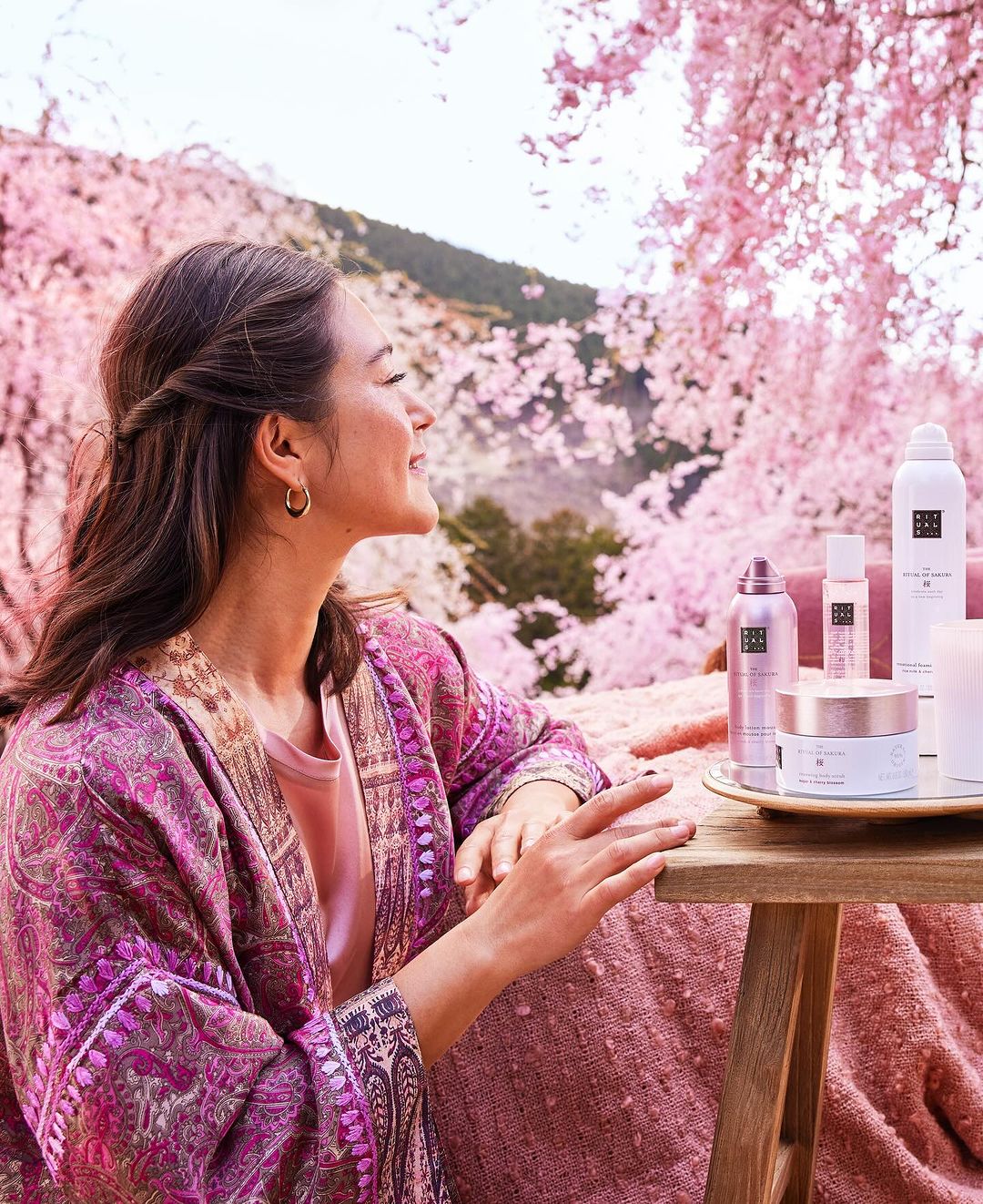 Woman in pink kimono sits outside with Rituals brand beauty products surrounded by pink cherry blossom trees in an outdoor setting.