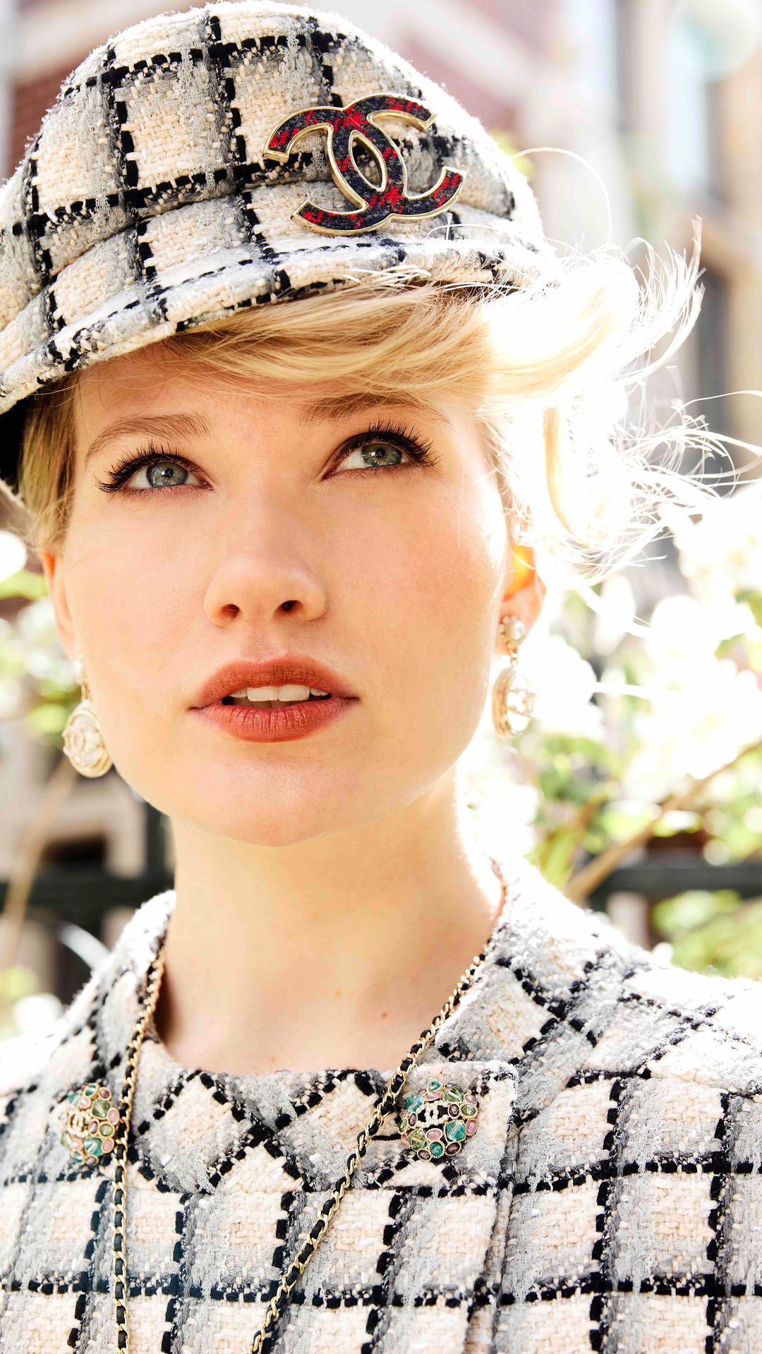 Blonde woman wearing Chanel tweed hat and jacket looking up with a Chanel logo necklace and earrings in an outdoor setting.
