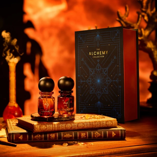 Two small brown glass perfume bottles with black spherical caps sit on old books, next to a black and blue decorative Alchemy Collection box.