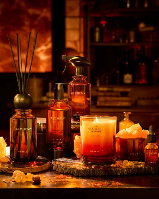 Still life of Rituals Alchemy collection featuring bottles, a candle, and decorative rocks on a dark surface with a dark background.