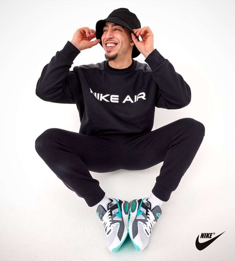 Man in black Nike tracksuit and bucket hat smiles while sitting cross-legged on white backdrop, displaying grey, white, and teal sneakers.
