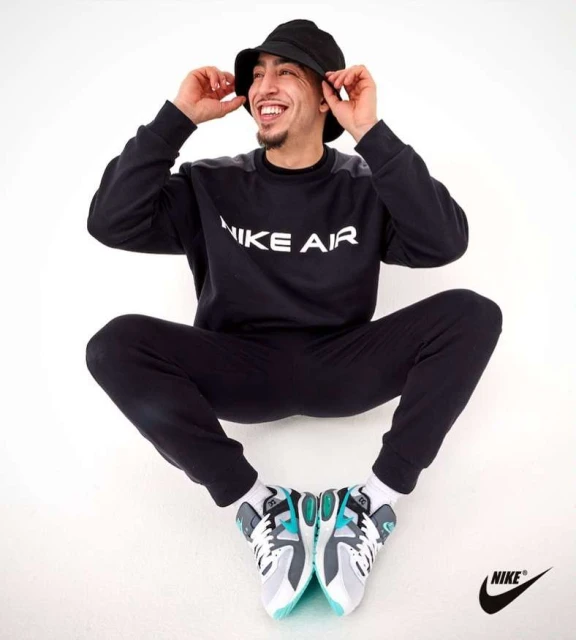 Man in black Nike tracksuit and bucket hat smiles while sitting cross-legged on white backdrop, displaying grey, white, and teal sneakers.