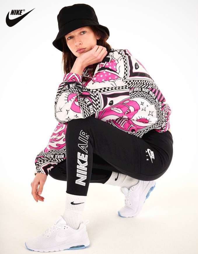 Model wearing a black bucket hat, patterned jacket, black leggings, and white sneakers crouching against a white background